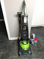 Full view of Bissell carpet steamer upright with hose and cleaning tool attached, standing on floor alongside cleaning solution bottles.