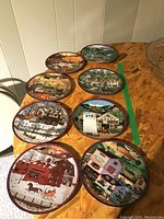 Photo showing 8 collector plates arranged on a table, colorful farm and rural scenes by Charles Wysocki, with deep maroon borders. Plates appear in very good condition.
