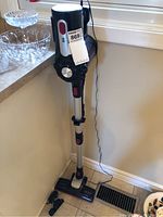 Full view of RoomieTEC cordless vacuum standing on floor next to windowsill, showing vacuum upright with power adapter connected.