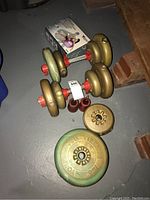 Photo of multiple dumbbells with gold and green weight plates, red collars, and red body ball weights on a floor next to bricks.