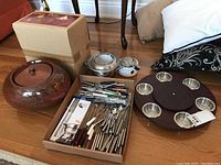 Photo of ceramic fire pot with lid next to original box, cardboard box containing many fondue forks and spoons, and wooden lazy susan with metal bowls.