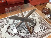 Overhead view showing the square beveled glass top with the unique wooden X-shaped base beneath it on a patterned area rug.