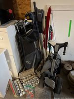 Photo showing Wilson branded black and gray golf bag filled with assorted mainly left handed golf clubs, mostly women's shorter clubs, alongside cartons of white golf balls and bags of orange golf tees on the ground.
