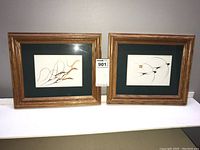 Two wooden framed Indigenous art prints with green matting displayed side by side on a white surface against a gray wall.