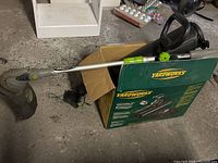 EarthWise weed wacker showing gray and green handle, black motor housing, curved shaft and spool head guard near end, laying on a cardboard box.