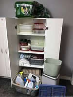 Photo of a white cabinet with open doors showing stacked plastic storage containers, folded towels, grey woven basket, green plastic garbage can, grey bathroom scale, and grey basket filled with assorted cleaning products on a dark floor.