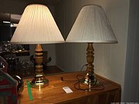 Pair of table lamps with brass-look circular bases and wooden turned bodies, topped with pleated fabric shades approximately 26 inches tall, placed on a wooden table.
