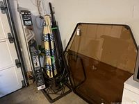 Large tinted glass patio table leaning against the wall with table frame visible, standing beside the folded patio umbrella in its stand.