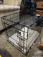 Front angle view of the black metal wire crate with Petmate brand sign and bedding pad inside, placed near couch and floor.