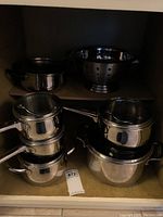 Four Lagostina pots stacked under cabinet with lids and a stainless steel colander on the shelf
