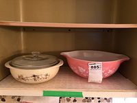 Two Pyrex bowls side by side on a shelf. Left is cream with brown floral pattern and glass lid, right is pink with white floral pattern.