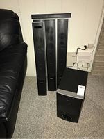 Four tall Sony SA-TS76W speakers standing upright and a Sony SS-WS78 subwoofer on carpet next to a black leather chair against a wall.