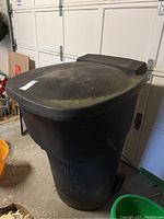 Front side view of large black plastic garbage bin with closed lid and two wheels on the bottom, showing wear and dust buildup.