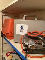 Side view of tools on shelf showing Black & Decker jig saw with orange cord and closed gray and orange carrying cases behind it.