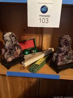 Photo showing two purple and white Chinese lion stone bookends on dark carved wooden bases, with a green patterned box, two green hand dexterity balls in a red-lined box, and two long packaged items (likely the fan and a fan box) on a wooden shelf.