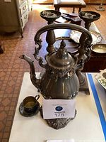 Wide view of antique silver coffee pot with curved handle, small cup and tray in front, and two tall candlesticks behind on a table.