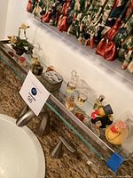 Various miniature perfume bottles, metal box, rubber duckies, and artificial flower decor arranged on glass shelf above sink with granite countertop and floral curtains in background.