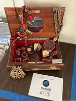 Wood jewelry box open showing red velvet lining and assortment of costume jewelry inside including brooches, earrings, chains, and a magnifying glass pendant.