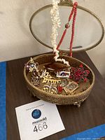 Full view of oval gold-tone metal jewelry box with glass top filled with assorted costume jewelry including necklaces and brooches.