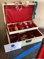 Photo of white multi-level jewelry box open showing red satin lining and various costume jewelry items organized in compartments and hanging on hooks
