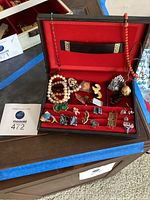 Black leather men's jewelry box opened, showing red velvet interior filled with assorted costume jewelry including pins, bracelets, and cufflinks.