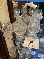 Shelf display showing one crystal decanter with round stopper and four matching brandy snifter glasses with intricate cut crystal patterns.