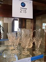 Crystal pitcher with four crystal glasses grouped together on a mirrored glass surface inside a wooden dresser cabinet.