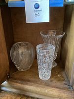 Three different crystal vases inside a wooden cabinet showing shape and size variation including round squat vase, tall cylindrical vase and a large fluted vase