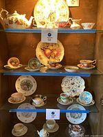 Overall photo showing multiple porcelain items including teacups, saucers, small dishes, and plates with fruit decoration arranged on glass shelves.