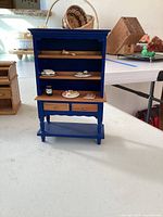 Miniature dark blue wooden hutch with two drawers and upper shelves holding tiny dishware and miniature food pieces on a white table, showing wear and chips on paint.