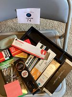 Photo shows various pencil boxes piled inside a black plastic desk organizer on a folding chair. Visible pencil boxes include Berol Mirado, Noir pens, Ticonderoga pencils, Universal pencils, and Verithin colored pencils. Small blue zipper case is also visible.