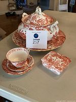 Full set showing soup tureen with lid, large serving tray underneath, teacup on saucer with small plate, and trinket box in red floral pattern with gold trim.