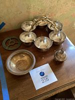 Photo showing six silver-tone bowls connected by brass handle with a dove, one separate silver-tone bowl, and Judaica items on wooden table