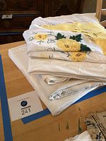 Stack of five folded tablecloths on wooden surface showing the variety of fabric, embroidery, and design including the yellow floral embroidery