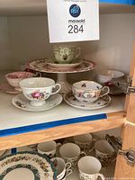 Multiple floral teacups with saucers and a pink floral cake plate displayed on shelf.