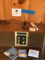 Photo of 1969 Heathkit GR-36 transistor radio in wooden cabinet with plug and antenna cable, three inspirational booklets, clear empty mezuzah case, and small steel brush displayed on wooden surface with wall behind. Blue tape showing dimensions 7x19x9 inches.