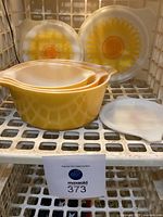 Photo showing three nested yellow Pyrex casserole dishes with lids and two large glass plates with large yellow and orange daisy designs placed behind them.