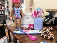 Overall view of American Girl surfer doll and accessories on wooden table including paddle, surfboard, suitcase, towel, necklaces, and pillows.