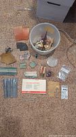 Full lot of assorted stones, wax wire pieces, jewelry making tools, files, and jump rings displayed on a carpeted floor with a bucket container.