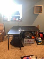 Photo showing two black folding tables of different sizes positioned in a room with carpeting and some clutter under and around them.