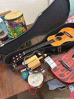 Photo showing two acoustic guitars, one in black case and one with red flames design, placed on a table with various percussion instruments including tambourine, wooden claves, maracas, and a small drum with rainbow strap