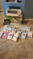 Cricut Expression machine with box, assorted Cricut cartridges laid out in front showing packaging and cable cords to the side
