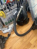 Black Electrolux canister vacuum with hose and metal wand on wooden floor near a baby gate and other household items