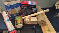 Photo showing the Honeywell fan box, Coolaroo shade sail bag, rolled shelf liner, IKEA wood boards, and other assorted home goods items on a table.