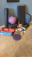 Photo showing assorted fitness items arranged on carpet: yoga balls, foam rollers, yoga mats, step platform, Bosu ball, DVDs, exercise cards, resistance bands, and hand massager.