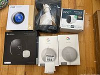 Photo of all items in the lot on a wooden floor including boxes of Nest Thermostat, Ecobee thermostat, 2 Google Nest Mini boxes, an Arlo camera box, and a Ring mount in bag.