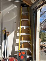 Full view of the yellow fiberglass ladder, showing side rails and metal steps, leaning against wall near other garage tools and objects.