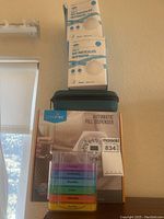 Photo of three boxes of N95 masks stacked on top of a Livefine automatic pill dispenser box, with pill organizer trays stacked in a clear plastic holder in front.