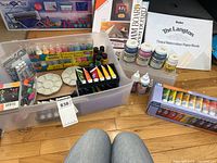 Overview of the lot showcasing a large plastic bin filled with assorted paints, paint bottles, a tinted watercolor paper book, a cardstock pack, and markers arranged around the bin on the floor.
