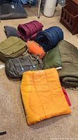 Photo of assorted sleeping bags in orange, green, blue, dark red, and folded or rolled on carpet.